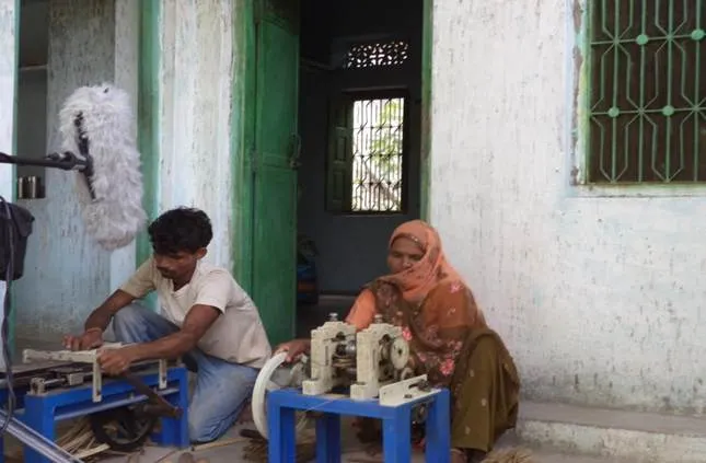 A man works a bamboo-processing machine on a blue metal stand while a woman sits at a smaller machine beside him, outside a house with a green door.