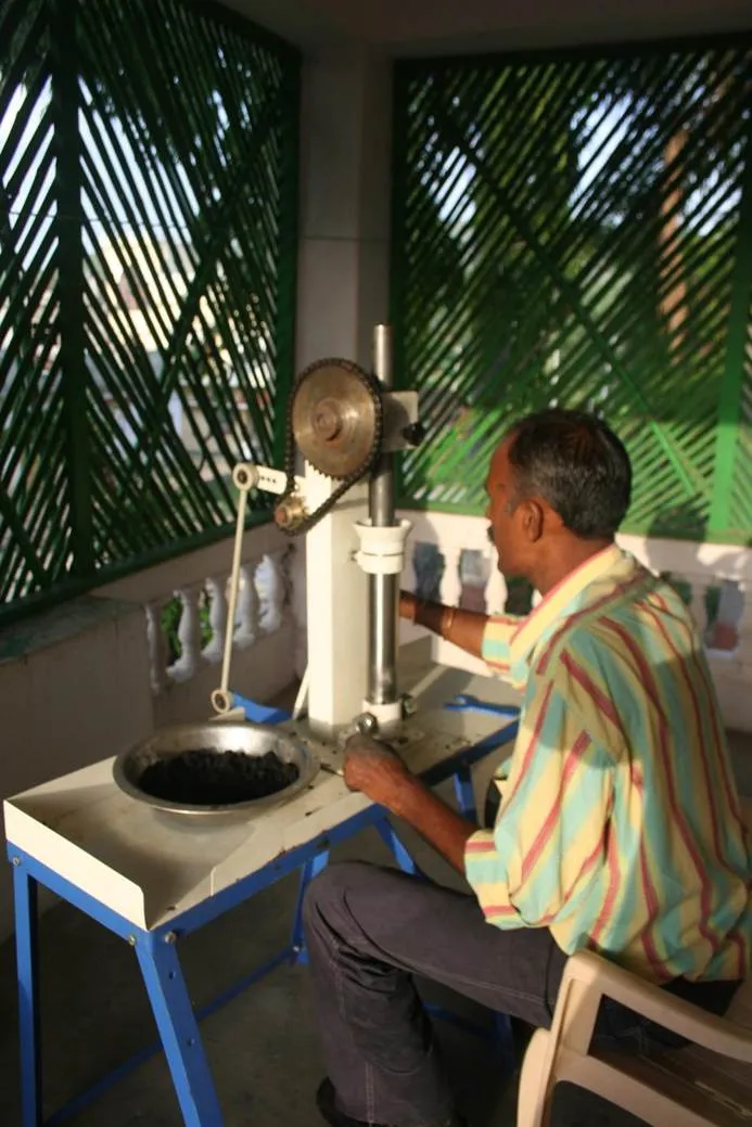 A man sits at an incense-rolling machine with a chain-and-sprocket drive, a steel bowl of dark incense mixture on the stand in front of him and green lattice screens behind.
